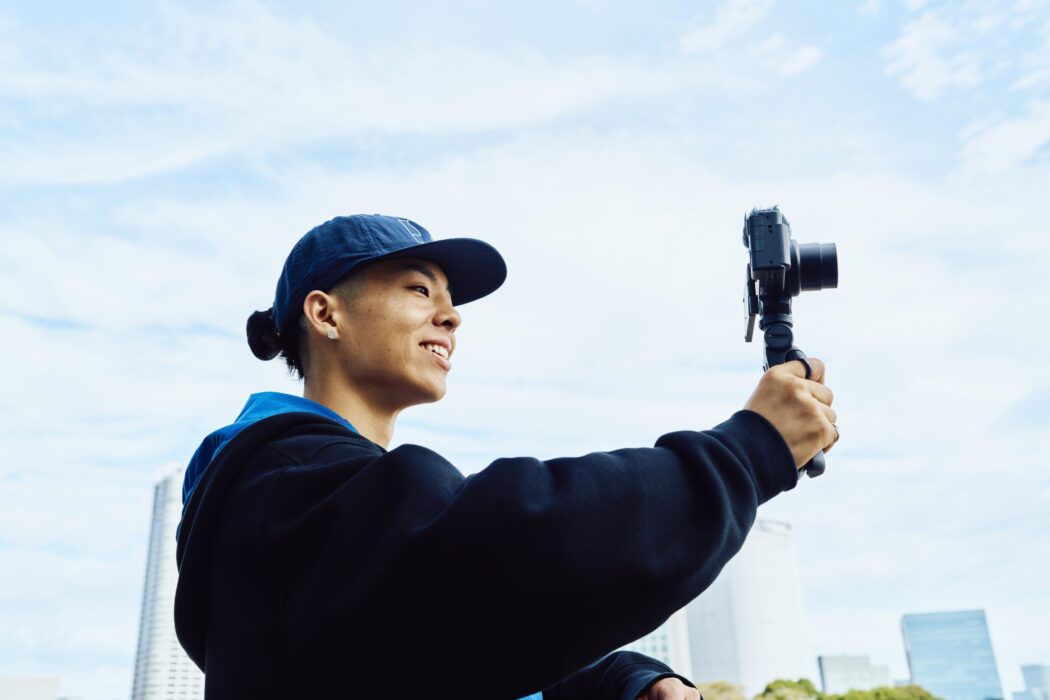 Uomo sorridente con cappellino che tiene in mano una fotocamera su un treppiede. Dietro, cielo azzurro e palazzi.