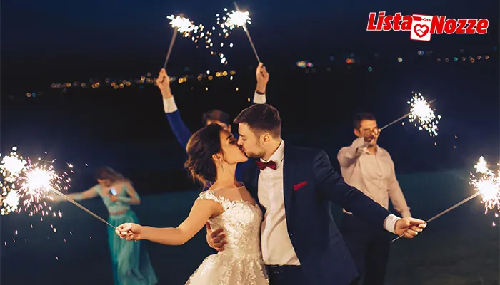 Coppia vestita elegante si bacia, circondata da persone con in mano fuochi d'artificio su sfondo scuro.