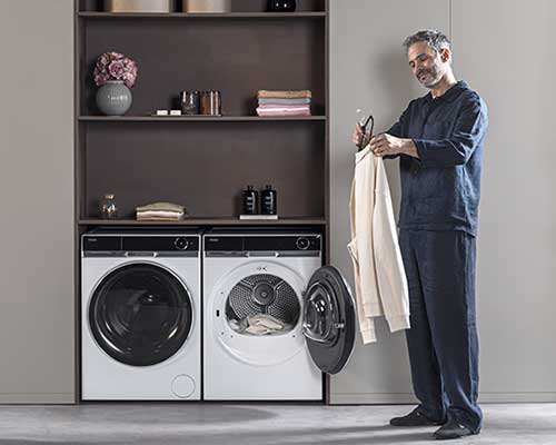 Uomo in lavanderia con lavatrice e asciugatrice bianche. Ripiani con vasi, asciugamani e flaconi. L'uomo tiene una camicia.