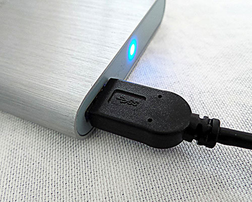 Cavo USB nero collegato a un disco rigido esterno grigio. Una luce blu � visibile sul dispositivo. Lo sfondo � bianco.