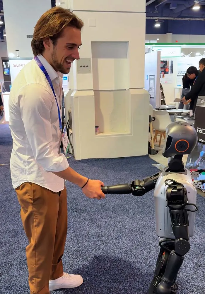 Uomo in camicia bianca stringe la mano a un robot grigio e nero su moquette blu in uno stand fieristico.
