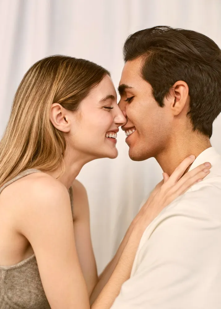 Donna in canotta e uomo in camicia bianca si toccano il naso e sorridono. La donna tiene la mano sulla spalla dell'uomo.