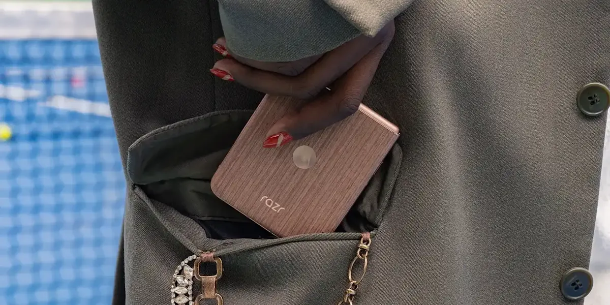Una mano con unghie rosse infila un telefono Razr marrone nella tasca di una giacca verde oliva. Sullo sfondo, una rete blu.
