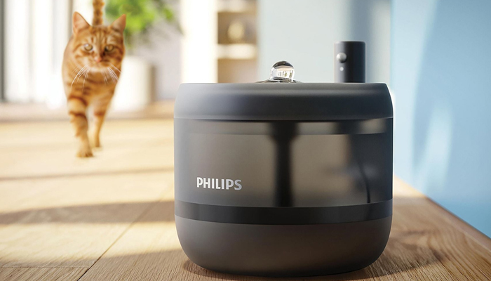 Una fontanella smart per gatti della Philips in primo piano, con un getto d'acqua a bolla sulla sommità. Sullo sfondo, un gatto soriano arancione cammina verso il dispositivo su un pavimento in legno chiaro in un ambiente domestico luminoso.