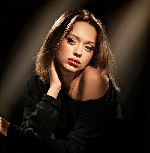 Donna con top nero su sfondo scuro illuminato. La mano destra è sollevata al collo.