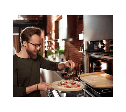 Egy szemüveges férfi pizzát készít a sütő közelében a konyhában. A pizza egy fahéjon van. Egy sütőlap van a sütőben.