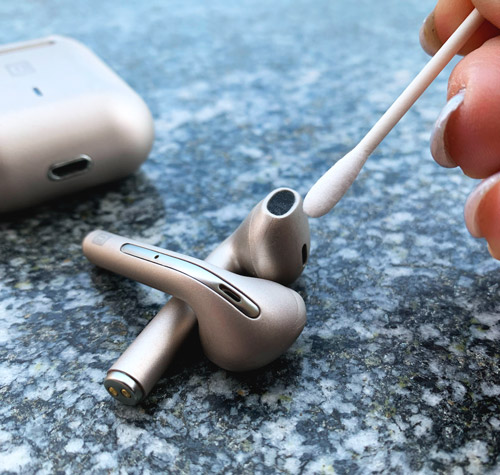 Auricolari e custodia bianchi su superficie marmorizzata. Una mano usa un cotton fioc per pulire gli auricolari.