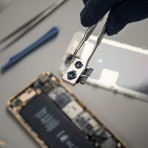 Mano guantata tiene pinzette con pezzo di elettronica sopra telefono smontato. Strumenti sul tavolo.