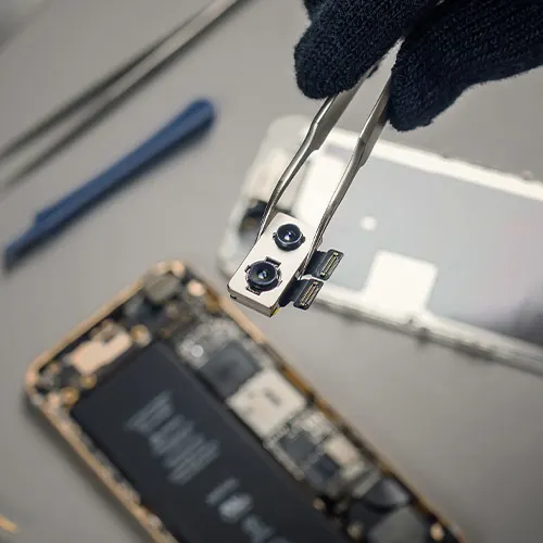 Mano guantata tiene pinzette con pezzo di elettronica sopra telefono smontato. Strumenti sul tavolo.