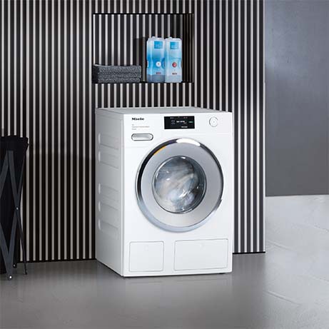 Lavatrice bianca Miele con obl� rotondo su pavimento grigio. Mensola con detersivi e parete a strisce verticali sullo sfondo.