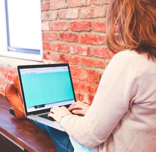 Donna che usa un laptop su una panca di legno con muro di mattoni sullo sfondo.