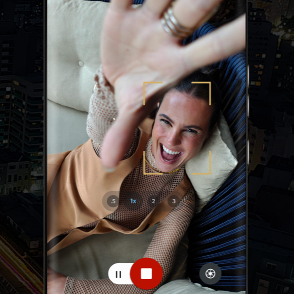 Donna sorridente su divano blu, ripresa da cellulare con interfaccia app. Mano alzata, sfondo notturno sfocato.