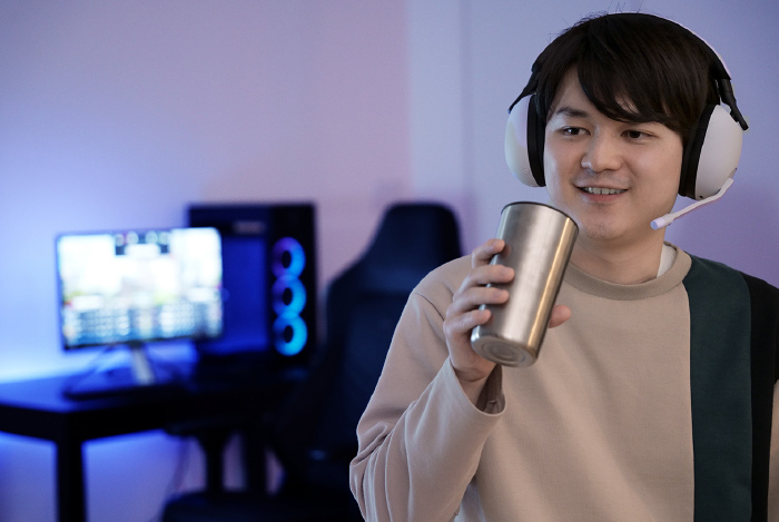 Uomo con cuffie che beve da un bicchiere di metallo. Dietro, schermo e computer da gaming su una scrivania nera.