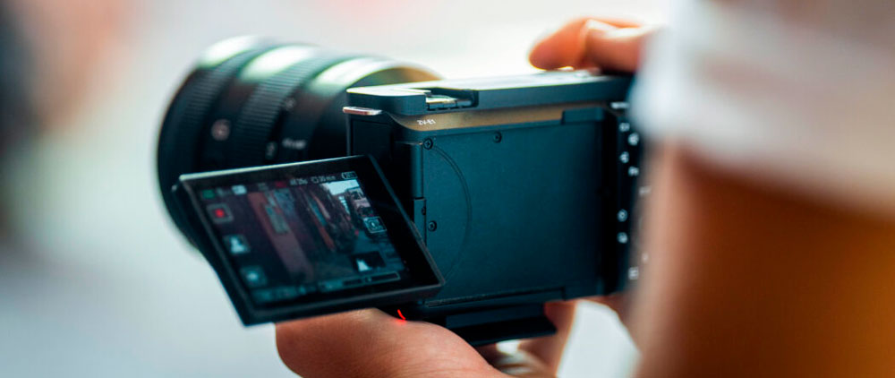 Una persona tiene in mano una fotocamera digitale nera con l'obiettivo puntato verso l'esterno e uno schermo orientabile aperto.