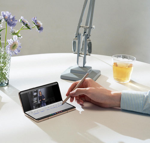 Su un tavolo bianco si trovano un tablet aperto con stilo, un bicchiere di birra, una lampada e un vaso di fiori.