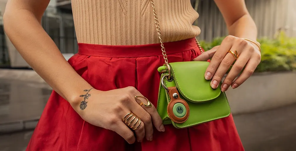Donna con gonna rossa e top beige tiene una borsa verde con tracolla a catena dorata e un portachiavi. Ha anelli alle mani.