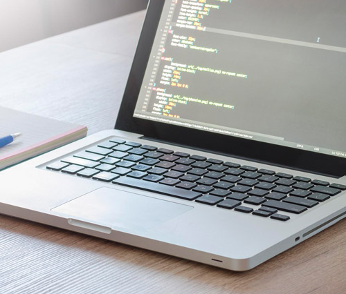Un computer portatile aperto su una scrivania mostra codice sullo schermo. Un quaderno e una penna sono accanto al portatile.