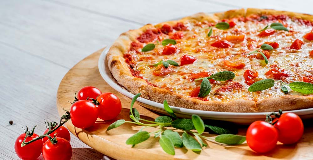 Pizza margherita su piatto con pomodorini e basilico su tagliere in legno. Sullo sfondo, tavolo bianco.