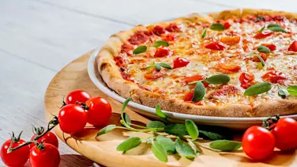 Pizza margherita su piatto con pomodorini e basilico su tagliere in legno. Sullo sfondo, tavolo bianco.