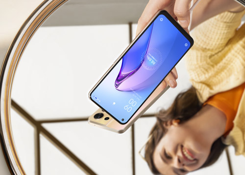 Riflesso in uno specchio: una mano tiene un telefono con schermo blu. Sullo sfondo, una figura sorridente indossa una camicia gialla.
