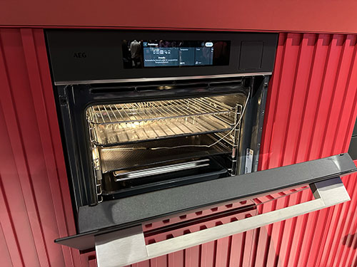 Forno nero aperto con griglie interne su parete rossa a listelli. Il pannello frontale è grigio e bianco.