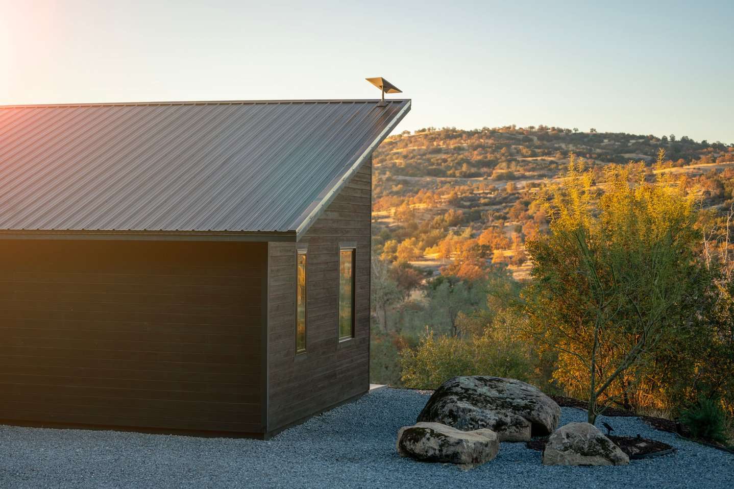 Casa moderna con tetto in metallo e pannello solare, su terreno di ghiaia. Dietro, colline boscose al tramonto.