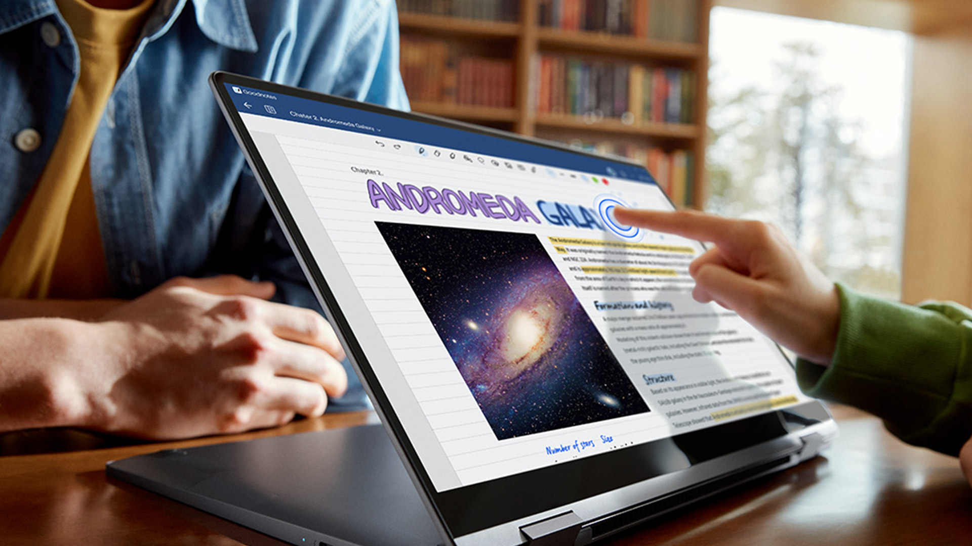 Su un tavolo di legno, un laptop aperto mostra una pagina con testo e un'immagine della galassia di Andromeda. Una mano indica lo schermo.