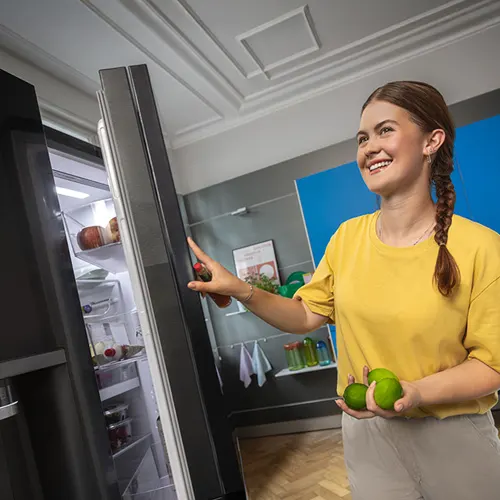 Donna in polo rossa indica un frigorifero aperto, con un uomo in primo piano e altri frigoriferi sullo sfondo.