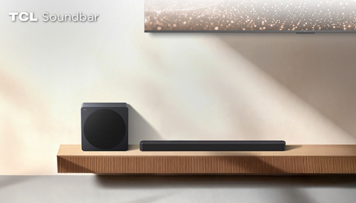 Una TCL Soundbar nera e il suo subwoofer wireless squadrato posizionati su una mensola in legno chiaro rigato, sotto la base di un televisore acceso.