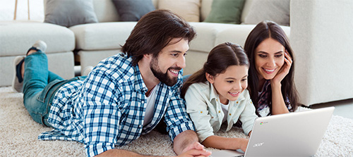 Una famiglia sorride guardando un laptop sul tappeto. L'uomo indossa una camicia a quadri blu e bianca. Divano chiaro sullo sfondo.