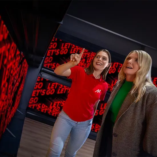 Due donne in interni con pareti rosse con scritta "GO LET'S GO". Una donna indica verso l'alto, l'altra indossa una giacca marrone.