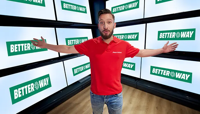 Uomo in polo rossa e jeans, braccia aperte. Sfondo di schermi multipli con scritta verde "Better Way" su sfondo bianco.