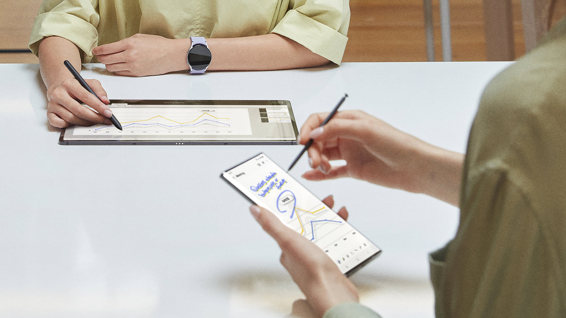 Persone che interagiscono con tablet e telefono con stilo su un tavolo bianco; grafici visualizzati.