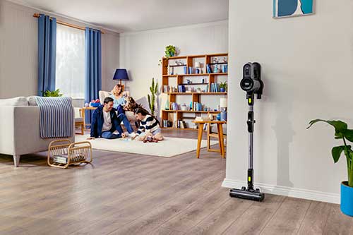 Interno di casa con famiglia seduta su tappeto, divano, libreria e aspirapolvere verticale grigio appoggiato al muro.