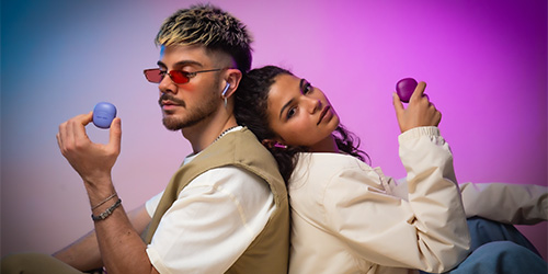 Uomo con occhiali da sole e gilet beige tiene un auricolare blu. Donna con maglia bianca tiene un auricolare viola.