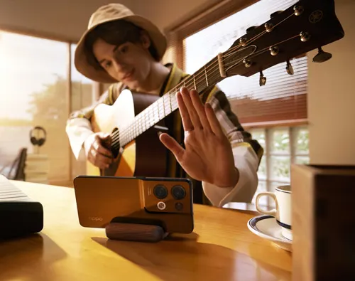 Un musicista con cappello suona la chitarra. Un telefono cellulare è posizionato sul tavolo. In sfondo una finestra.