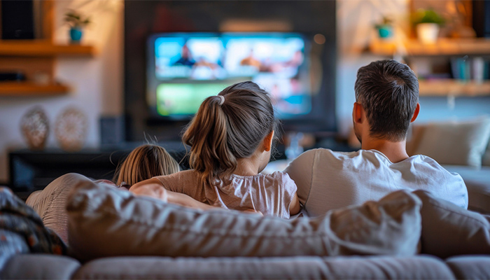 Padre e due bambini seduti sul divano guardano la TV. Scaffali di legno e piante sullo sfondo.
