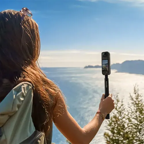 Ragazza riprende il mare con telecamera su asta. Il cielo � azzurro e l'acqua riflette la luce. Alberi visibili a destra.