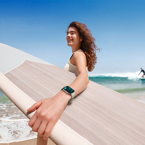 Donna sorridente con tavola da surf beige in spiaggia. Indossa un orologio al polso. Cielo blu e mare sullo sfondo.