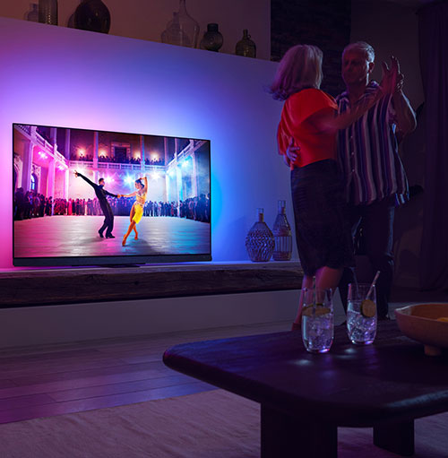 Due persone ballano in salotto davanti a una TV che mostra una coppia che balla in una sala da ballo. Sul tavolino ci sono due bicchieri.