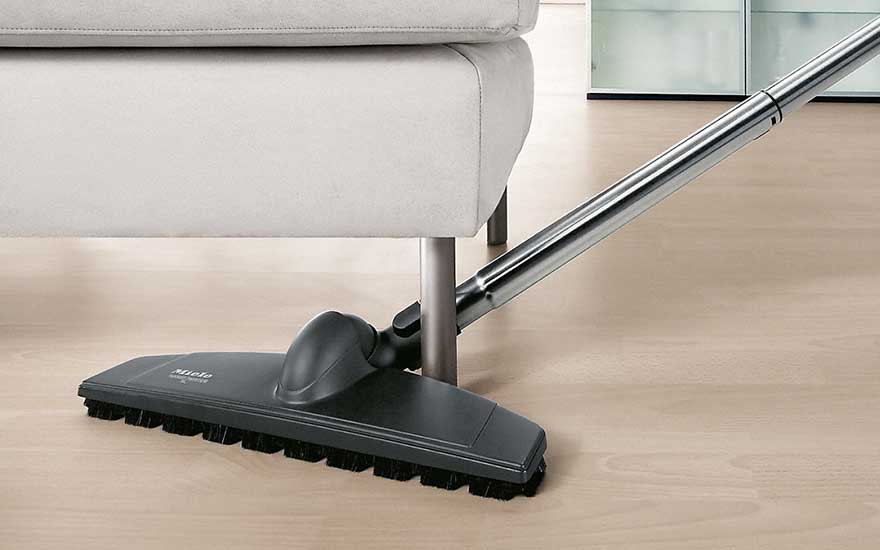 A vacuum cleaner easily cleans under a sofa with metal legs, on a wooden floor in a modern living room. 