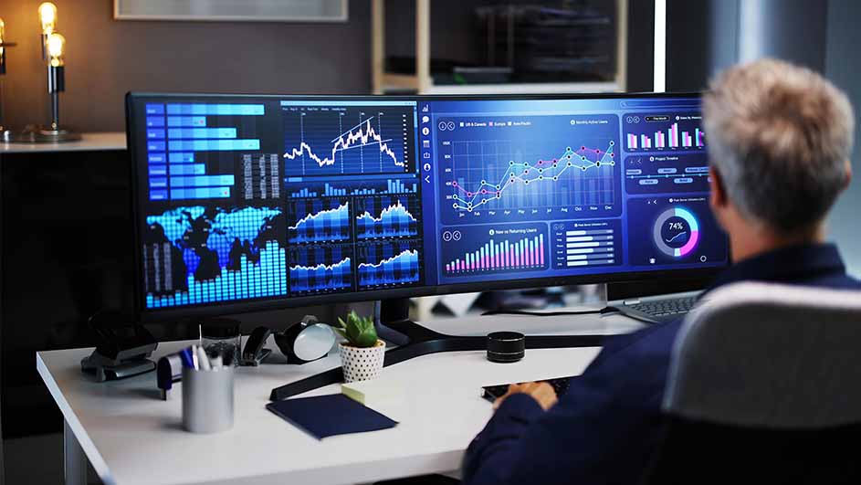 Een man zit achter een bureau met een breed beeldscherm dat blauwe grafieken en data weergeeft.