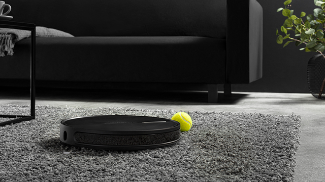 Zwarte robotstofzuiger op een grijs tapijt naast een gele tennisbal, met een zwarte bank en bijzettafel op de achtergrond.