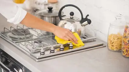 Een hand met een gele doek maakt een zilverkleurig gasfornuis schoon. Er staan pannen en potten op het aanrecht.