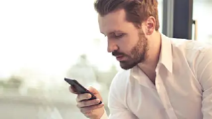 Een man in een wit overhemd kijkt naar een telefoon in zijn hand. Achter hem een raam en een kamerplant.