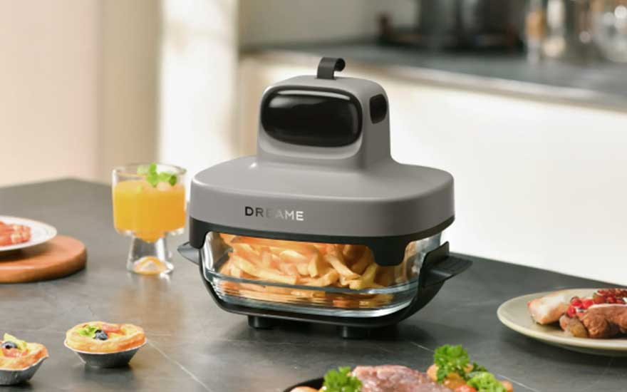 Een compacte Dreame airfryer met glazen bak bereidt frietjes op een keukenblad, omringd door drankjes en eten.