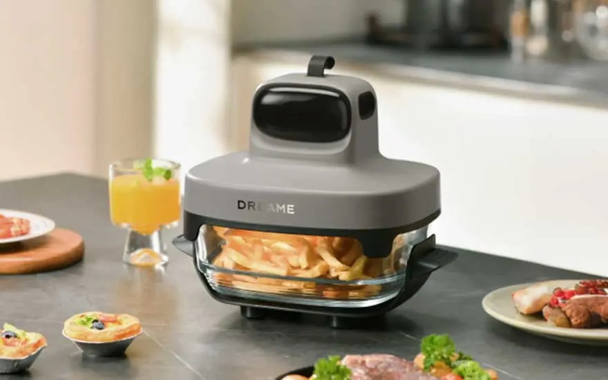 Een compacte Dreame airfryer met glazen bak bereidt frietjes op een keukenblad, omringd door drankjes en eten.