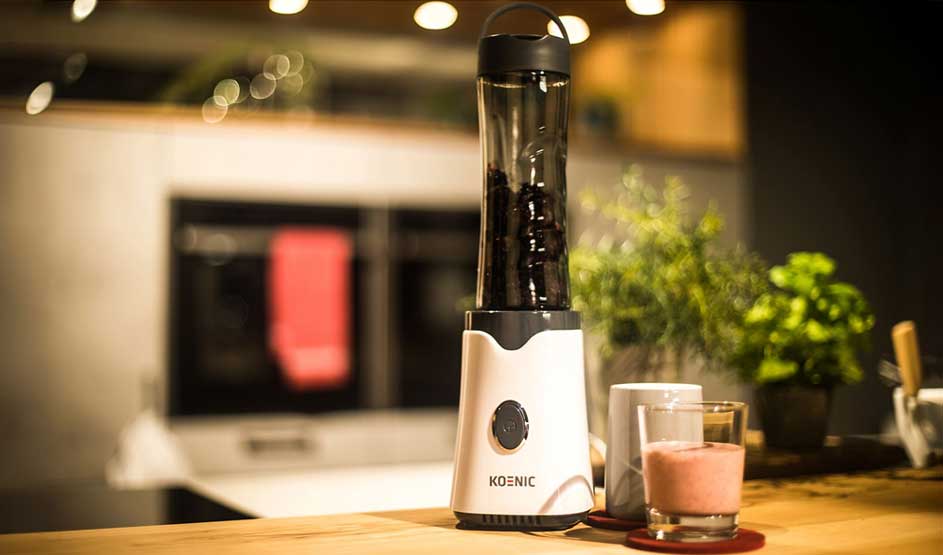 A slim blender stands on a wooden counter; in front a glass of pink smoothie and a mug, with herb plants and warm bokeh lights.