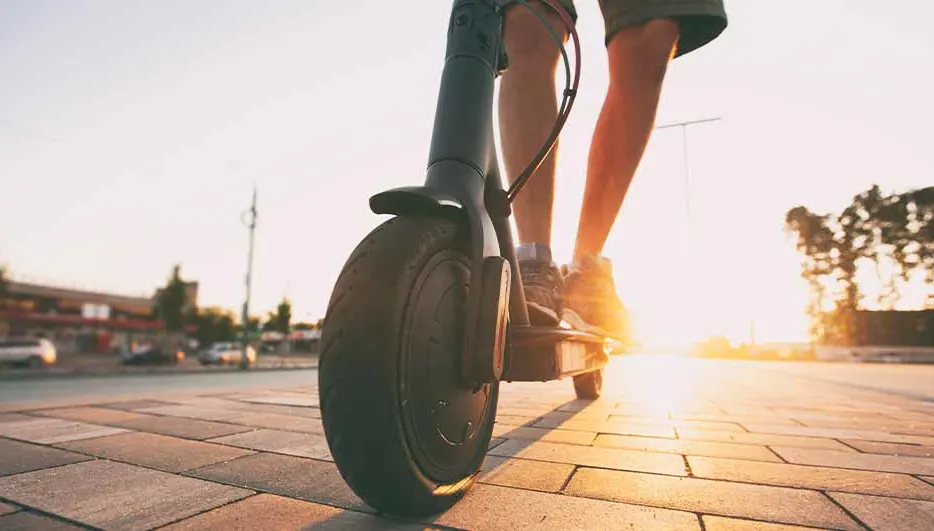 Een persoon rijdt op een grijze elektrische step op een bestrate weg bij zonsondergang, met gebouwen en bomen op de achtergrond.