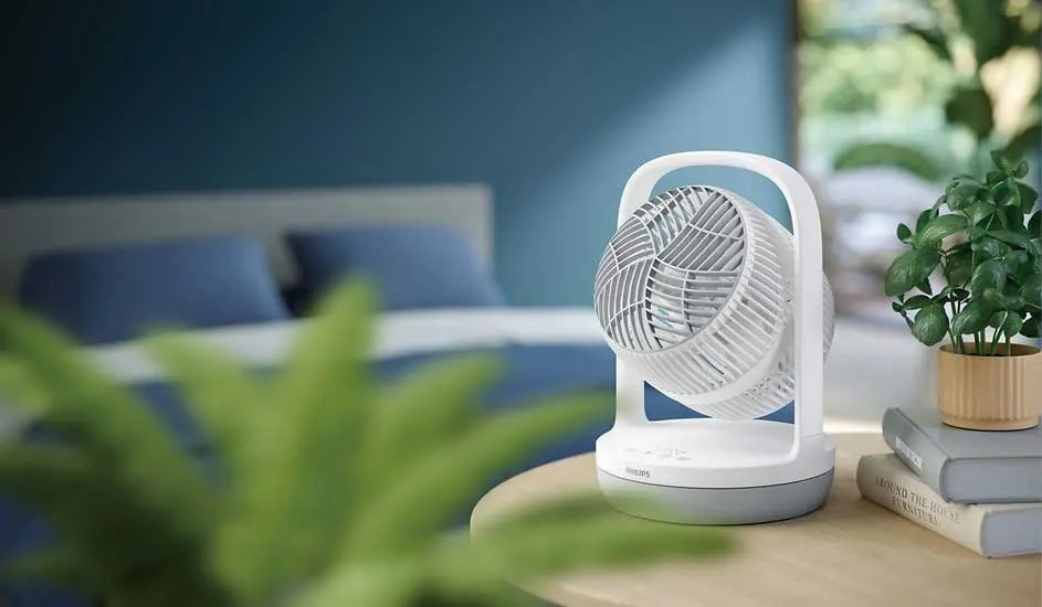 White table fan spins in a calm bedroom, surrounded by green plants and books on a small wooden bedside table.